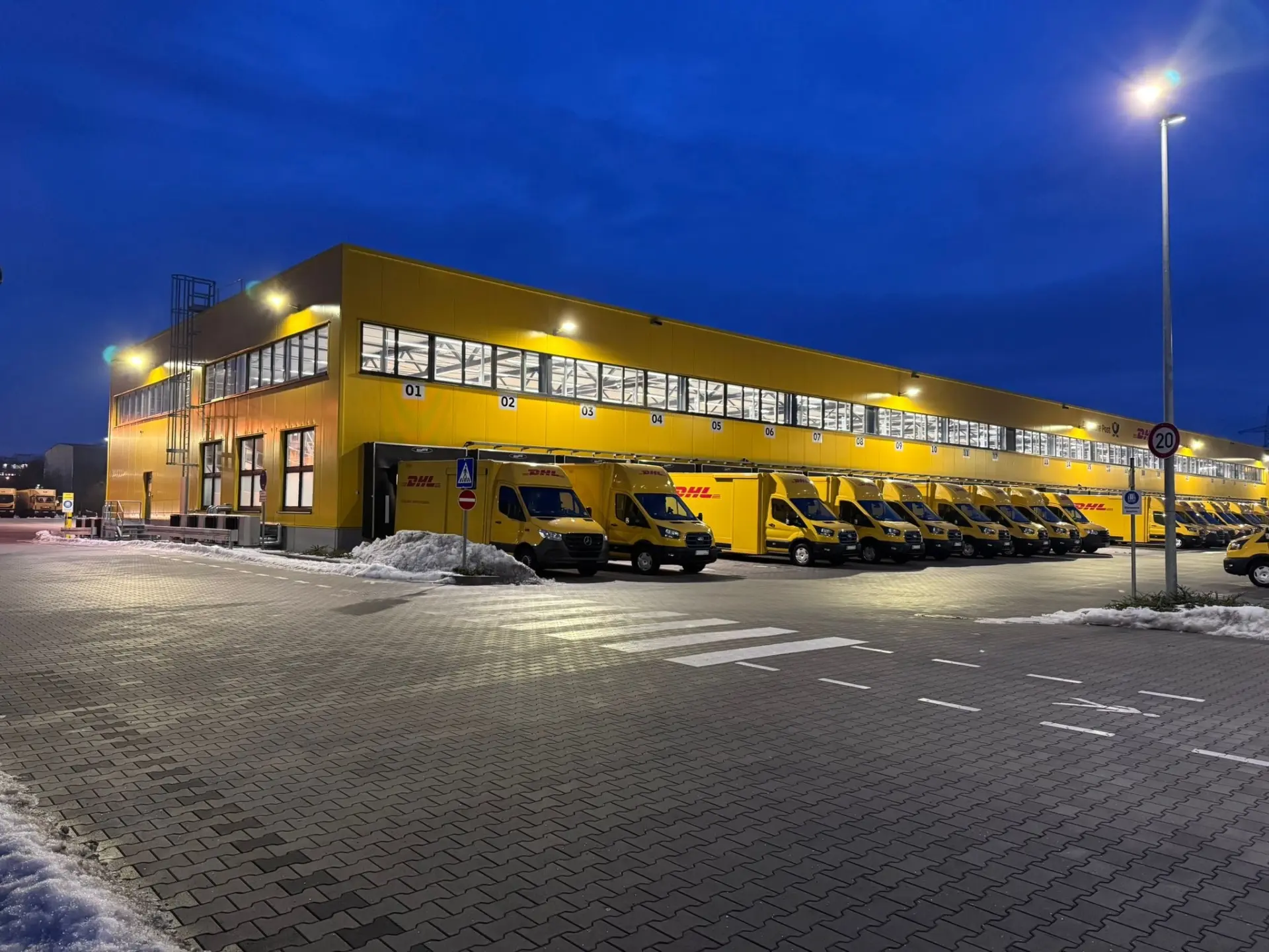 Neubau des Logistikstandorts DHL Regensburg in Regensburg bei Nacht mit beleuchteter Fassade und Transportern an den Laderampen.