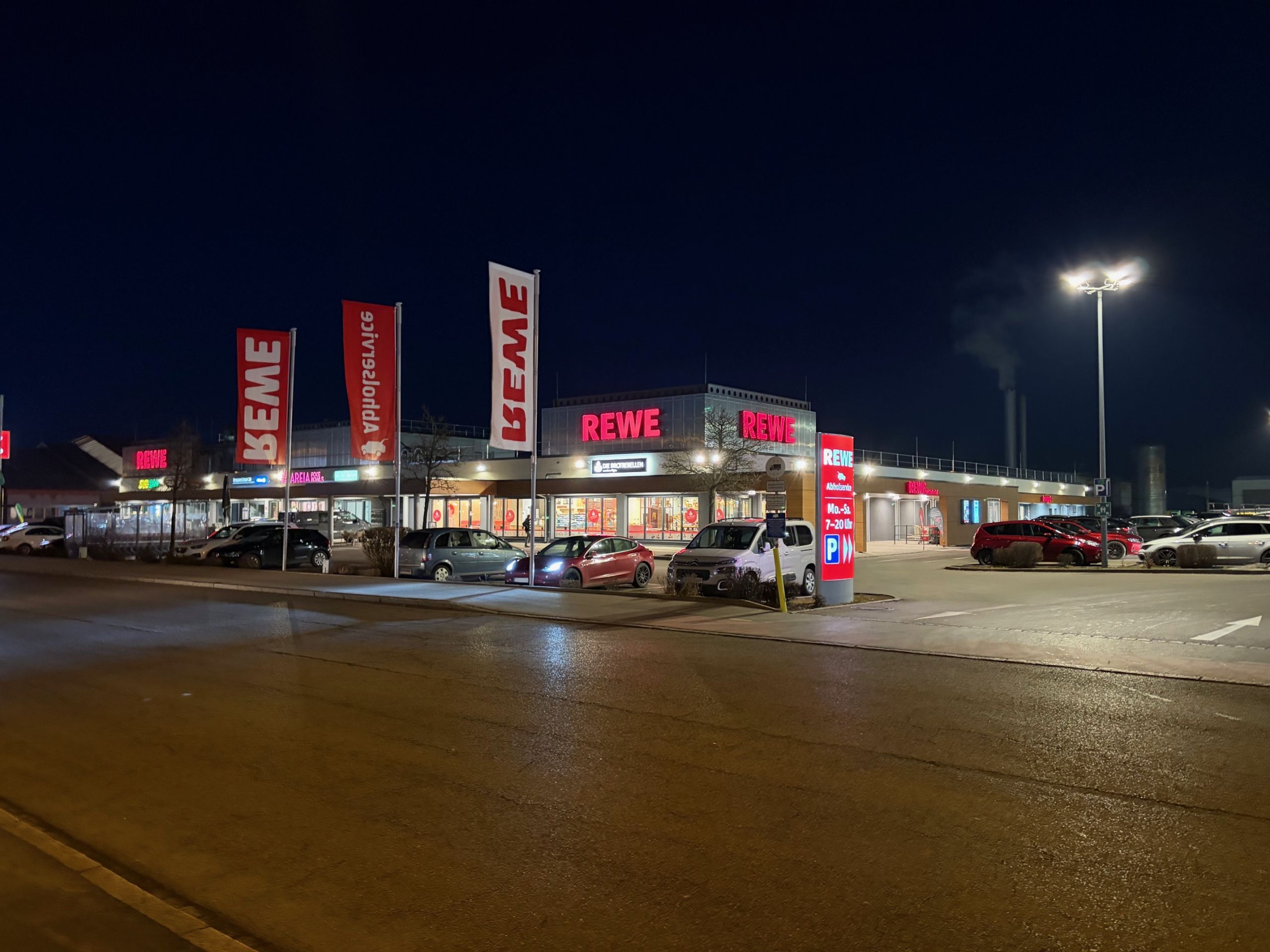 Außenansicht des neu gebauten Rewe-Marktes in Memmingen bei Nacht mit beleuchteter Fassade und Kundenparkplatz.